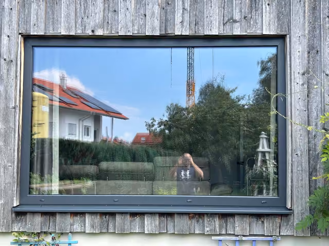 Großes Panoramafenster mit dunklem Rahmen in einer Fassade aus vergrauten Holzbrettern