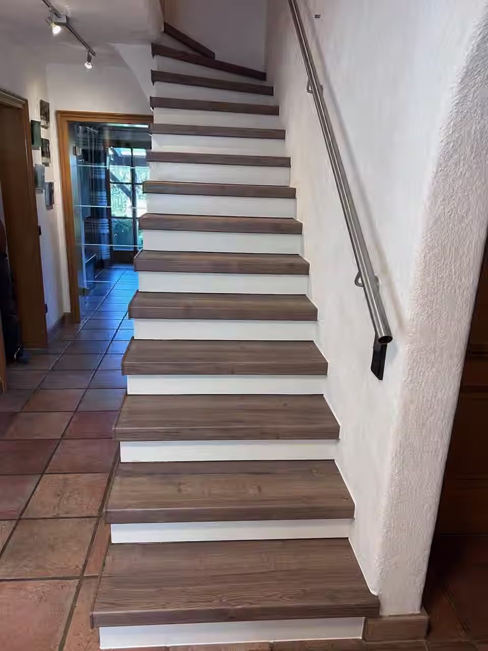 Treppe mit Eichen-Trittstufen und weißen Setzstufen über einem Terrakottaboden, Handlauf an der Wand